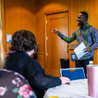 man smiling holding a paper, fellow presenter behind him, woman in flowered shirt watching